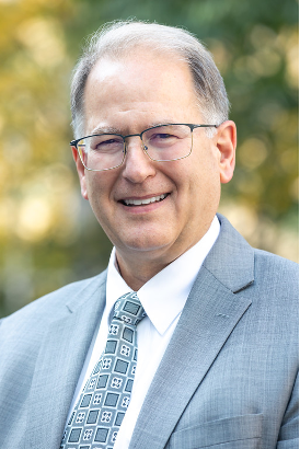 A man in professional attire and glasses smiles in an outdoor setting