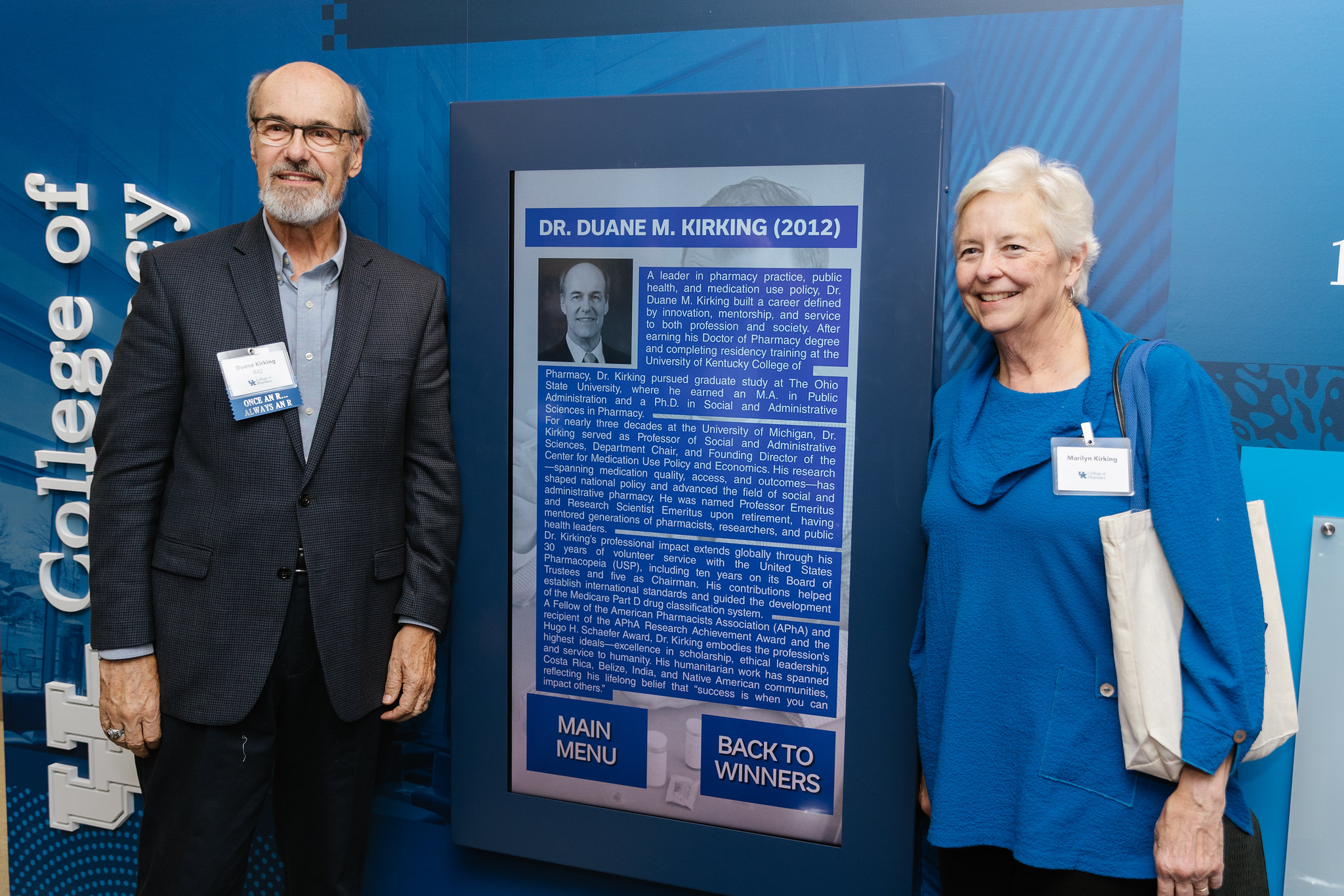 Two older adults, a man and woman, pose next to an interactive screen displaying the headshot and bio of Duane M. Kirking