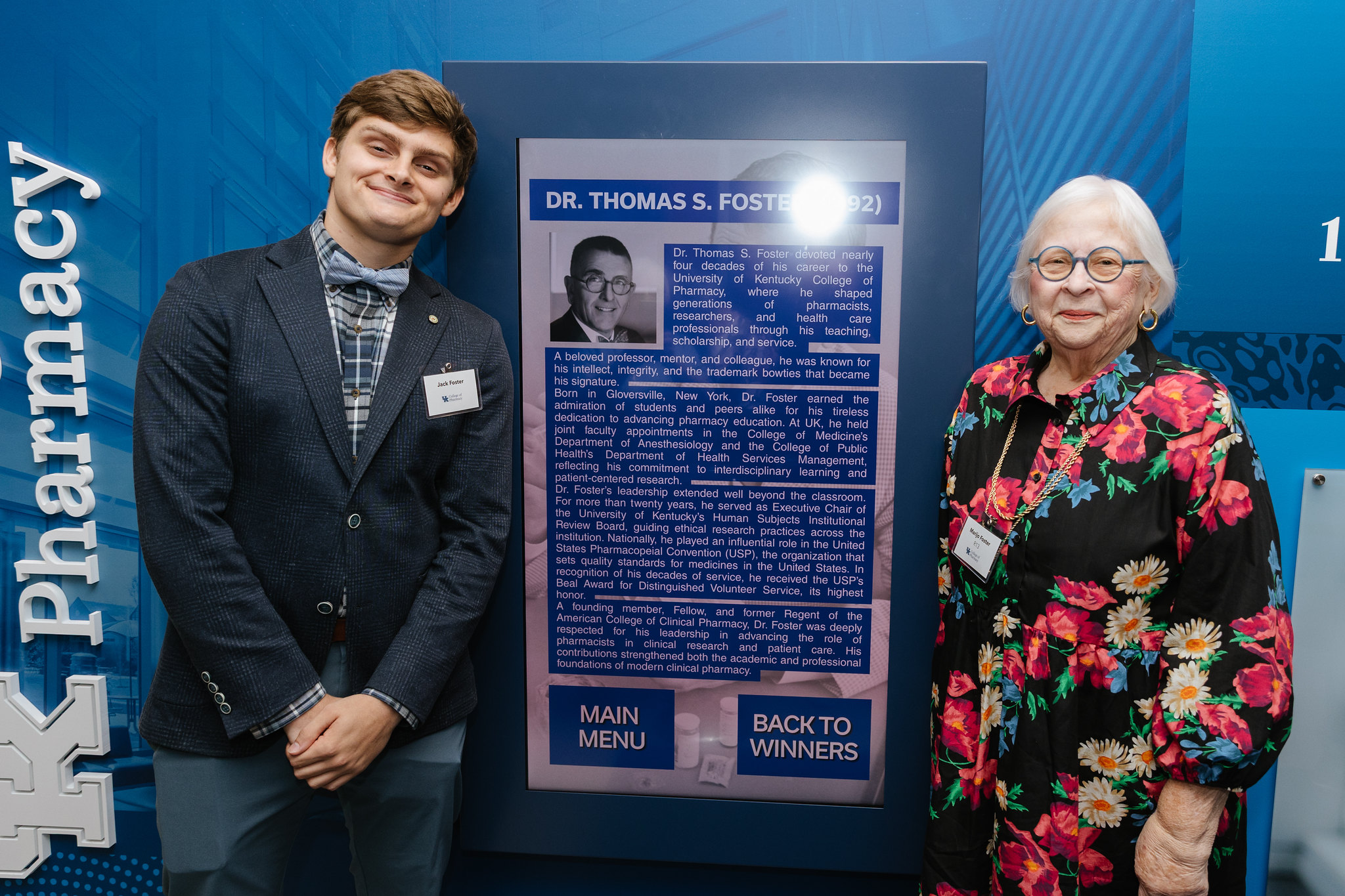 A young man and older adult woman pose next to an interactive screen with a photo and biography of Scott Foster