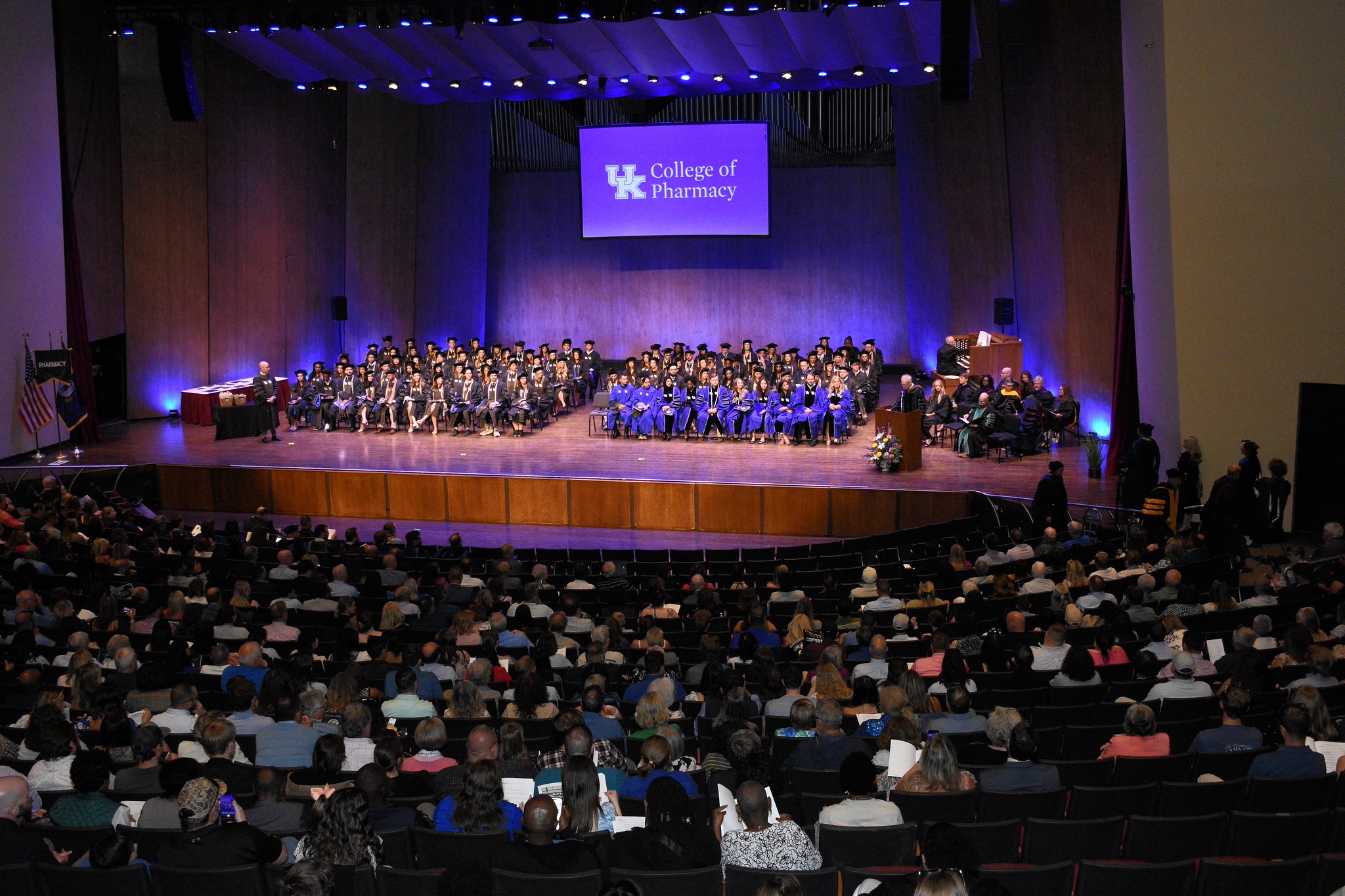 A view from the audience of a stage full of sitting graduates with the UK college of pharmacy logo shown on the screen behind them