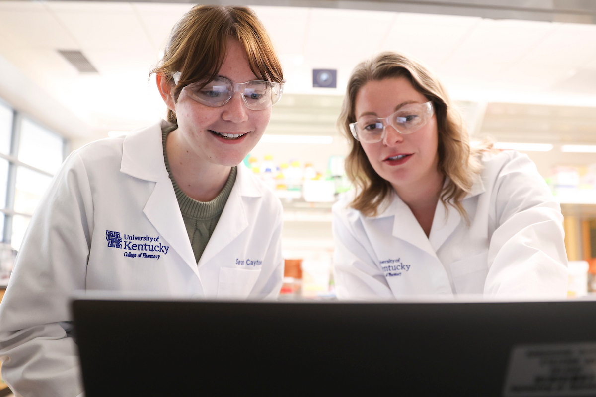two women in white coats look at a computer screen in a lab setting