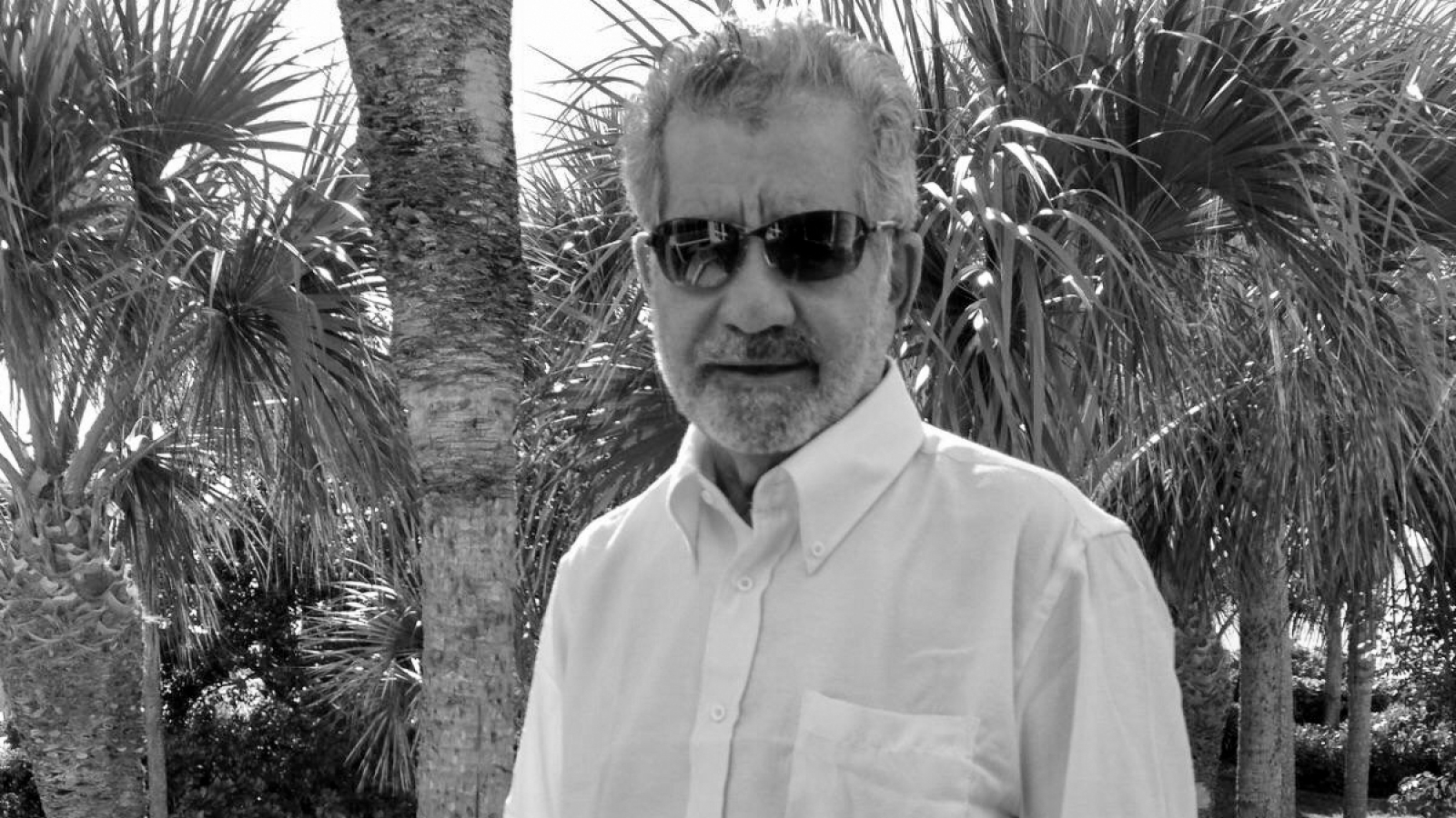 A black and white photo of a man standing outside, wearing a button down and sunglasses