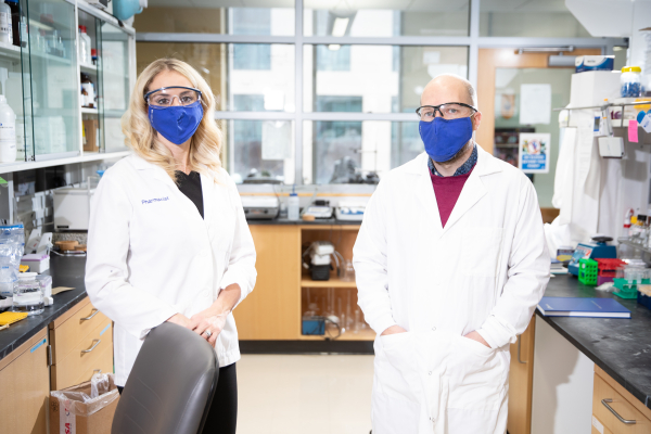 Brooke Hudspeth and Vince Venditto wearing white coats and protective eyewear while standing in a research lab