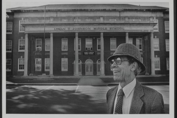Lyman T Johnson wearing a suite, hat and glasses posing in front of Lyman T. Johnson Middle School