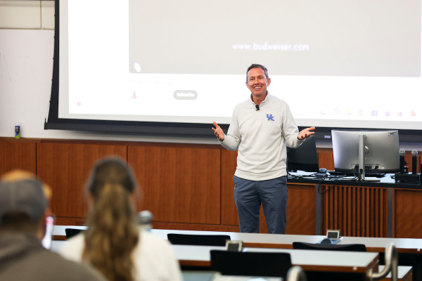Dr. Jeff Cain stands at the front of a lecture hall, smiling and gesturing openly with both hands while addressing students. He is wearing a gray University of Kentucky pullover. A projection screen is visible behind him.
