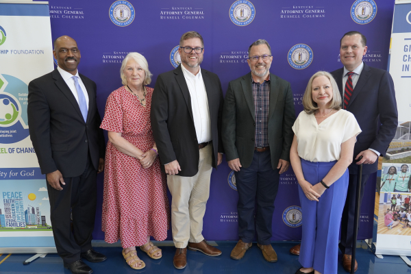 Six people stand smiling shoulder to shoulder in front of a purple backdrop bearing the seal of the Kentucky Attorney General's Office, at an event announcing opioid settlement research grants.
