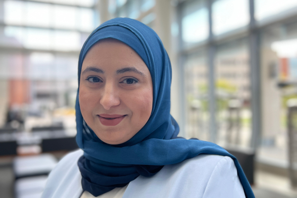 Professional headshot of Dr. Hend Mansoor smiling warmly at the camera. She is wearing a light blue blazer and a blue hijab. The background shows a bright, modern interior space with large windows.