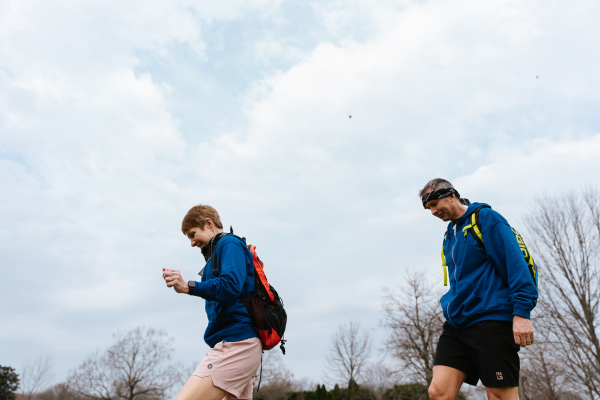 Two people wearing blue hoodies and backpacks walk outdoors on a trail under a cloudy sky, appearing focused as they move along a slightly elevated path with trees in the background.