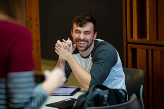smiling male student sitting at table with hands clasped