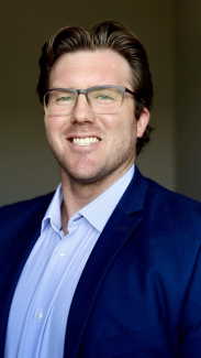 A man wearing a blazer, button down shit, and glasses smiles at the camera