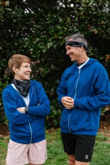 A woman and man in matching blue hoodies and black bandanas look at each other and smile