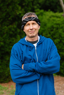 A man in a blue hoodie poses with arms crossed in an outdoor setting, smiling slightly