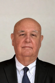 An older man in a black suit jacket and tie and white button down smiles pleasantly in front of a neutral background
