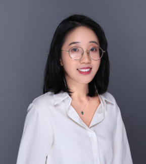 A dark haired woman wearing glasses and a white button down shirt smiles in front of a neutral backdrop