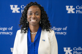 a woman in a blue shirt and white jacket stands in front of a blue uk college of pharmacy backdrop