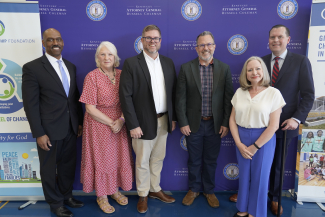 Six people stand smiling shoulder to shoulder in front of a purple backdrop bearing the seal of the Kentucky Attorney General's Office, at an event announcing opioid settlement research grants.
