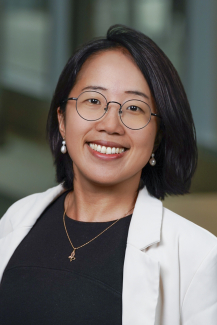 A woman with short hair wearing glasses and a white suit jacket smiles in an indoor setting