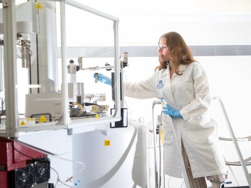 person on step stool standing in white coat toward an NMR machince