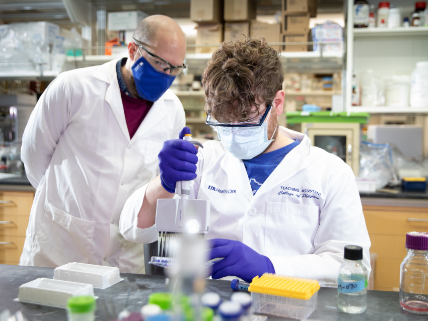 Two scientists in research lab wearing masks and white coats, one looks over the others shoulder