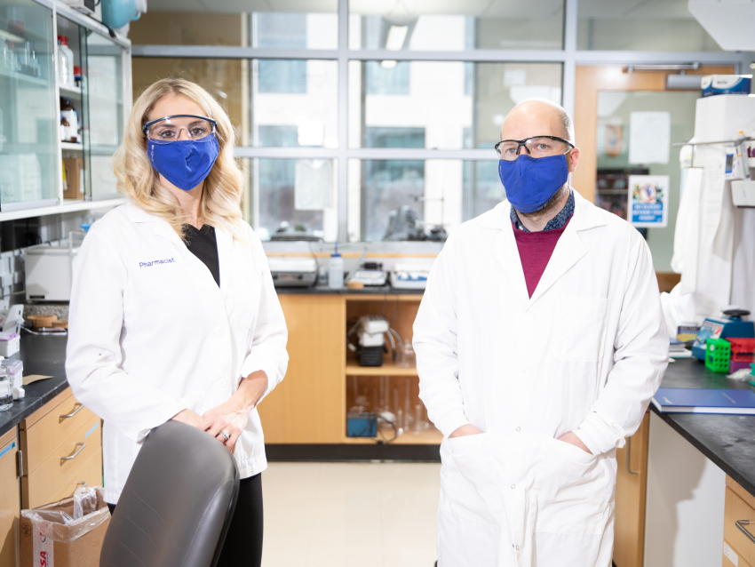 Brooke Hudspeth and Vince Venditto wearing white coats and protective eyewear while standing in a research lab