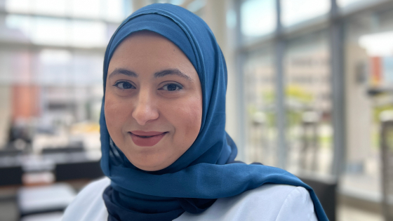 Professional headshot of Dr. Hend Mansoor smiling warmly at the camera. She is wearing a light blue blazer and a blue hijab. The background shows a bright, modern interior space with large windows.