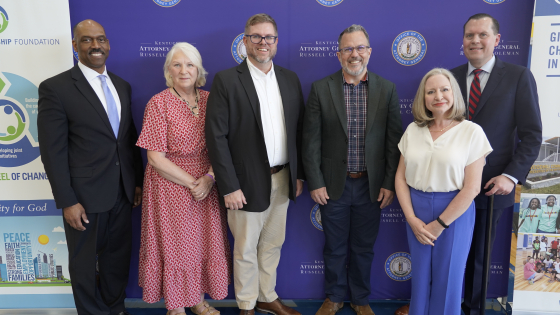 Six people stand smiling shoulder to shoulder in front of a purple backdrop bearing the seal of the Kentucky Attorney General's Office, at an event announcing opioid settlement research grants.