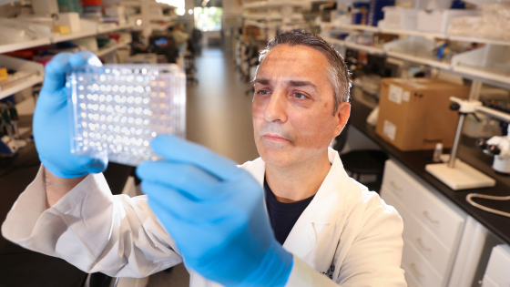 A man in white coat and lab gloves observes equipment held up to the light
