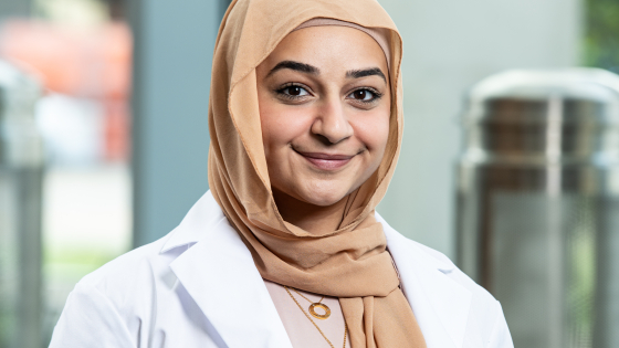 A young woman in a hijab and white coat with the UK College of pharmacy logo smiles at the camera