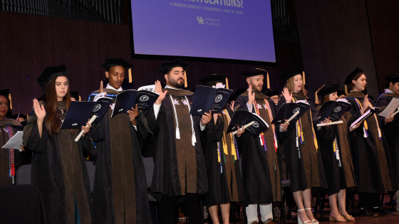 Graduates on stage in full regalia with their right hand in the air, taking the oath of a pharmacist