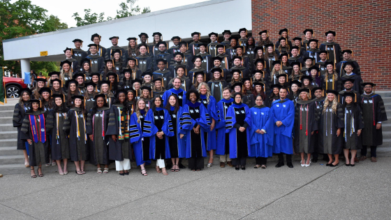 A large group of graduates wearing doctoral realia pose on stairs in an outdoors setting