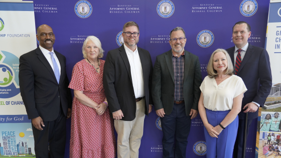 Six people stand smiling shoulder to shoulder in front of a purple backdrop bearing the seal of the Kentucky Attorney General's Office, at an event announcing opioid settlement research grants.