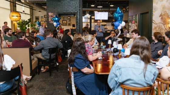 A group of people sit at dining tables inside a restaurant while someone speaks into a microphone in the background