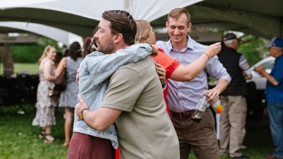 Four people hugging one another in an outdoor setting. A large white tent and more people visible in the back.