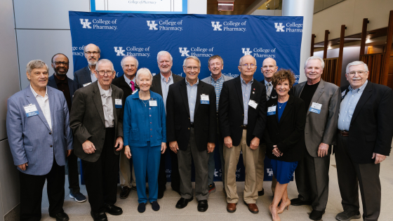 a group of older adults wearing business casual attire stand in front of a photo backdrop with the UK College of Pharmacy logo on it