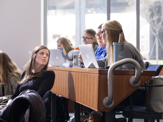 group of students sitting at a desk with laptops, with one students in front leaning back talking