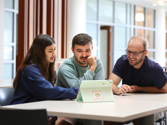 three students sitting at a table looking at an tablet