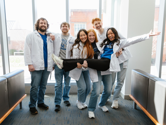 Group of six smiling students in white pharmacists coats standing in a bright hallway, with four of them holding a laughing classmate horizontally in front of them.