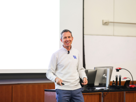 A person smiling at the front of a classroom setting, facing the class
