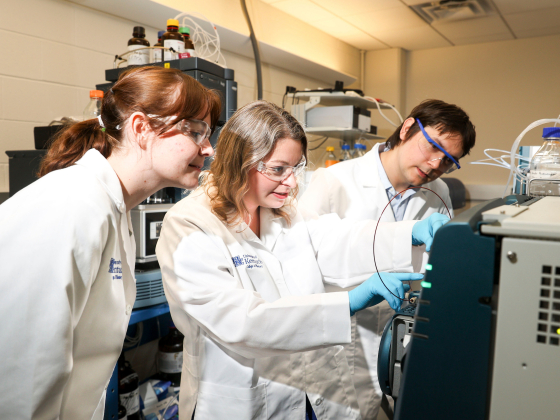 three people in white coats work on equipment in a lab
