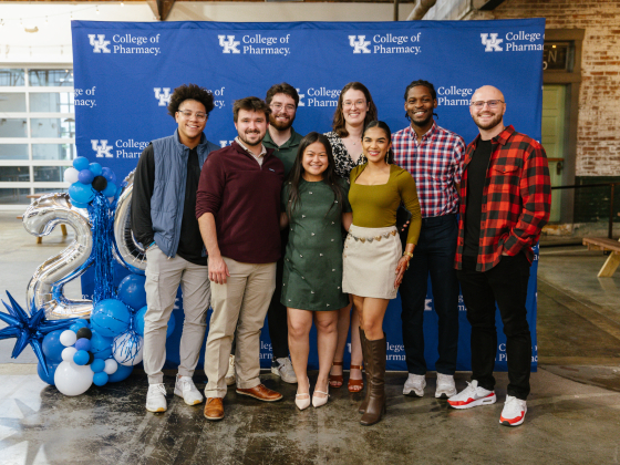 A group of students pose in front of a blue UK College of Pharmacy backdrop