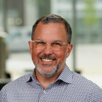 A man wearing a button down and clear frame glasses smiling