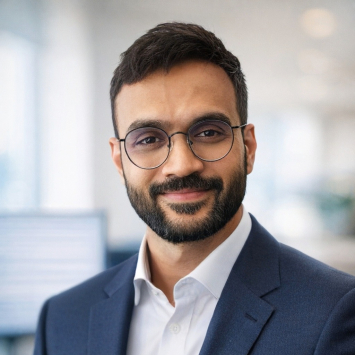 A dark haired bearded man wearing a white button down, navy blazer and glasses smiles at the camera