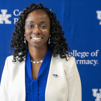 a woman in a blue shirt and white jacket stands in front of a blue uk college of pharmacy backdrop