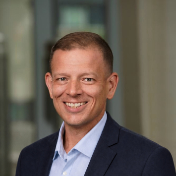 A man in a blue button down and navy jacket smiles at the camera 