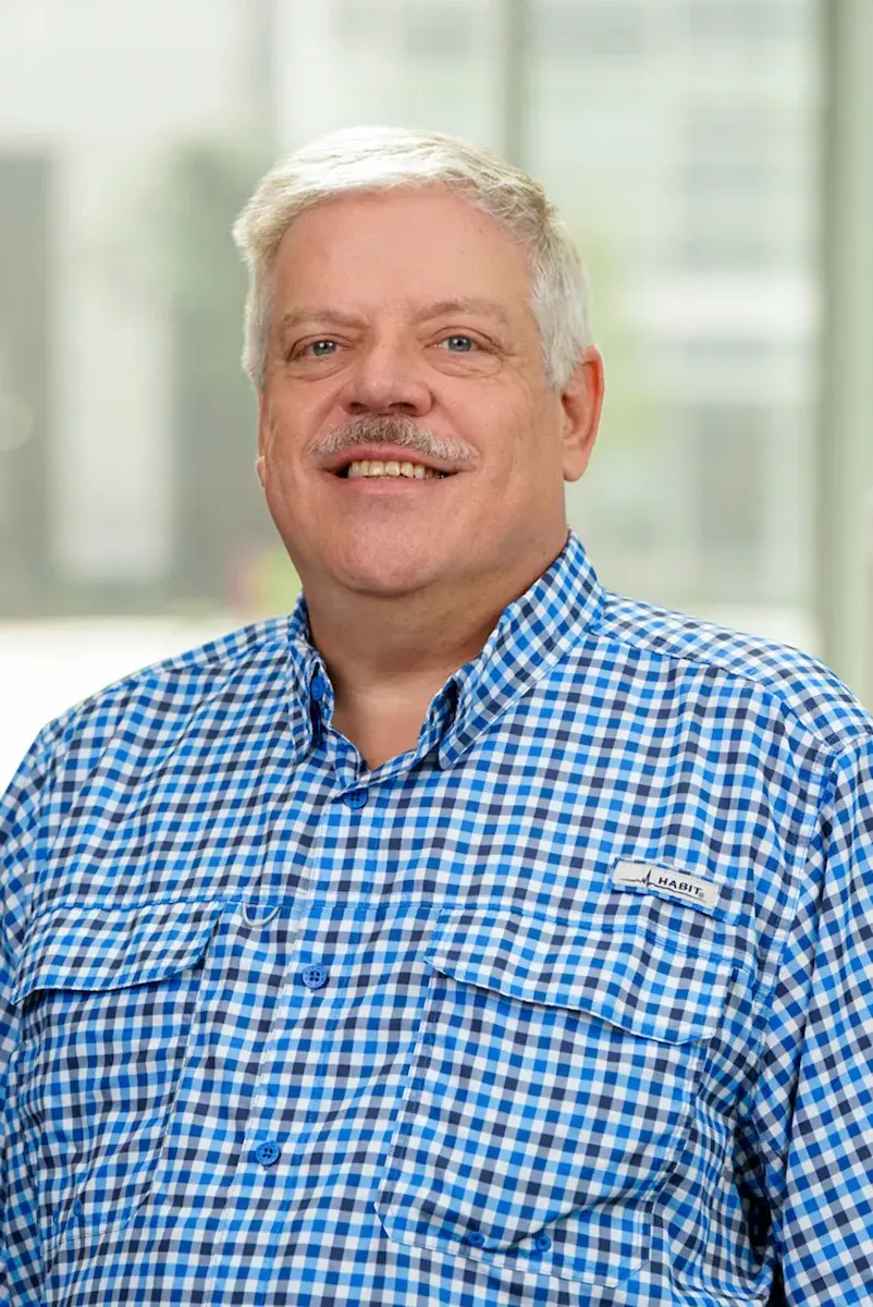 A gray haired mustached man wearing a button down shirt smiling