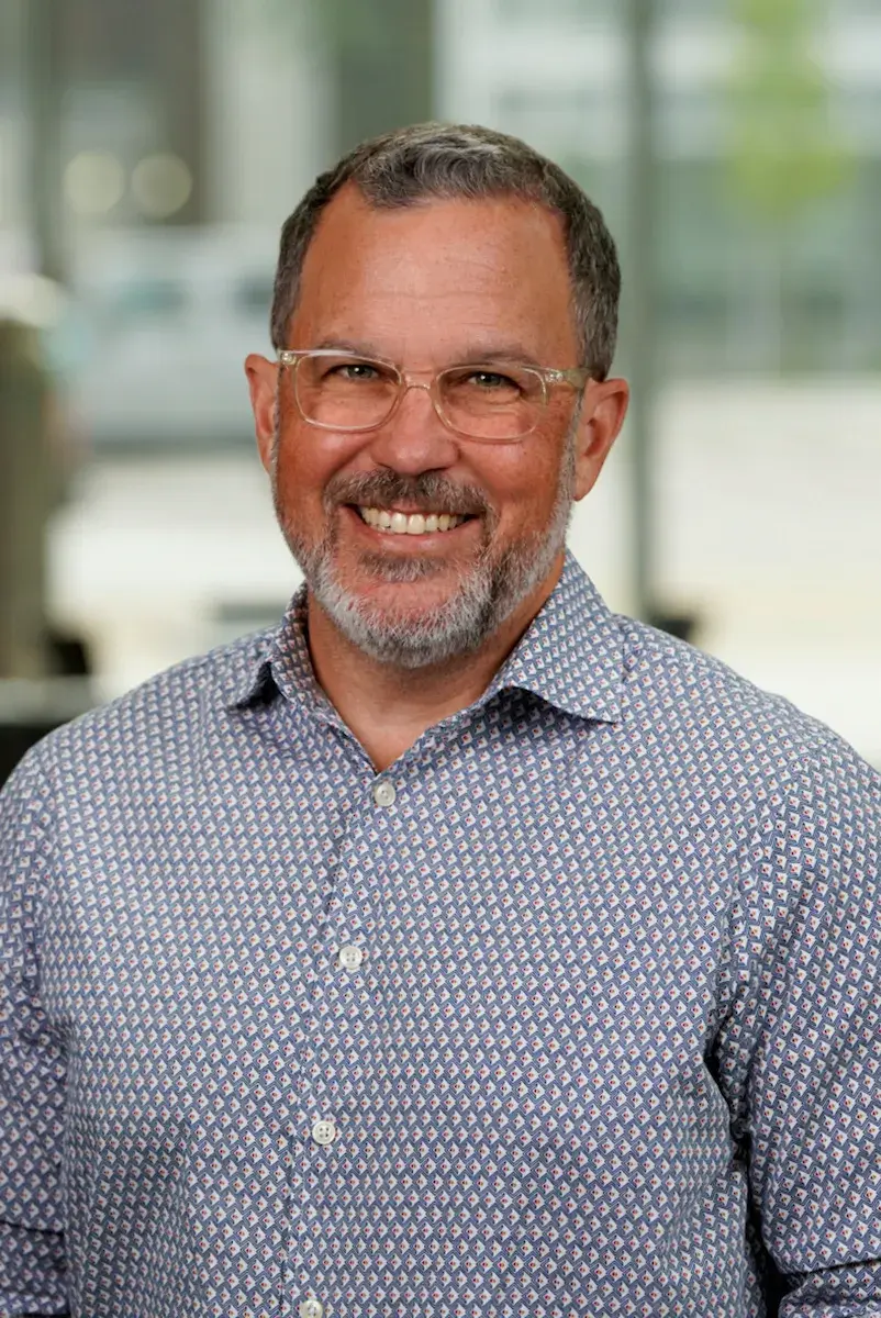 A man wearing a button down and clear frame glasses smiling