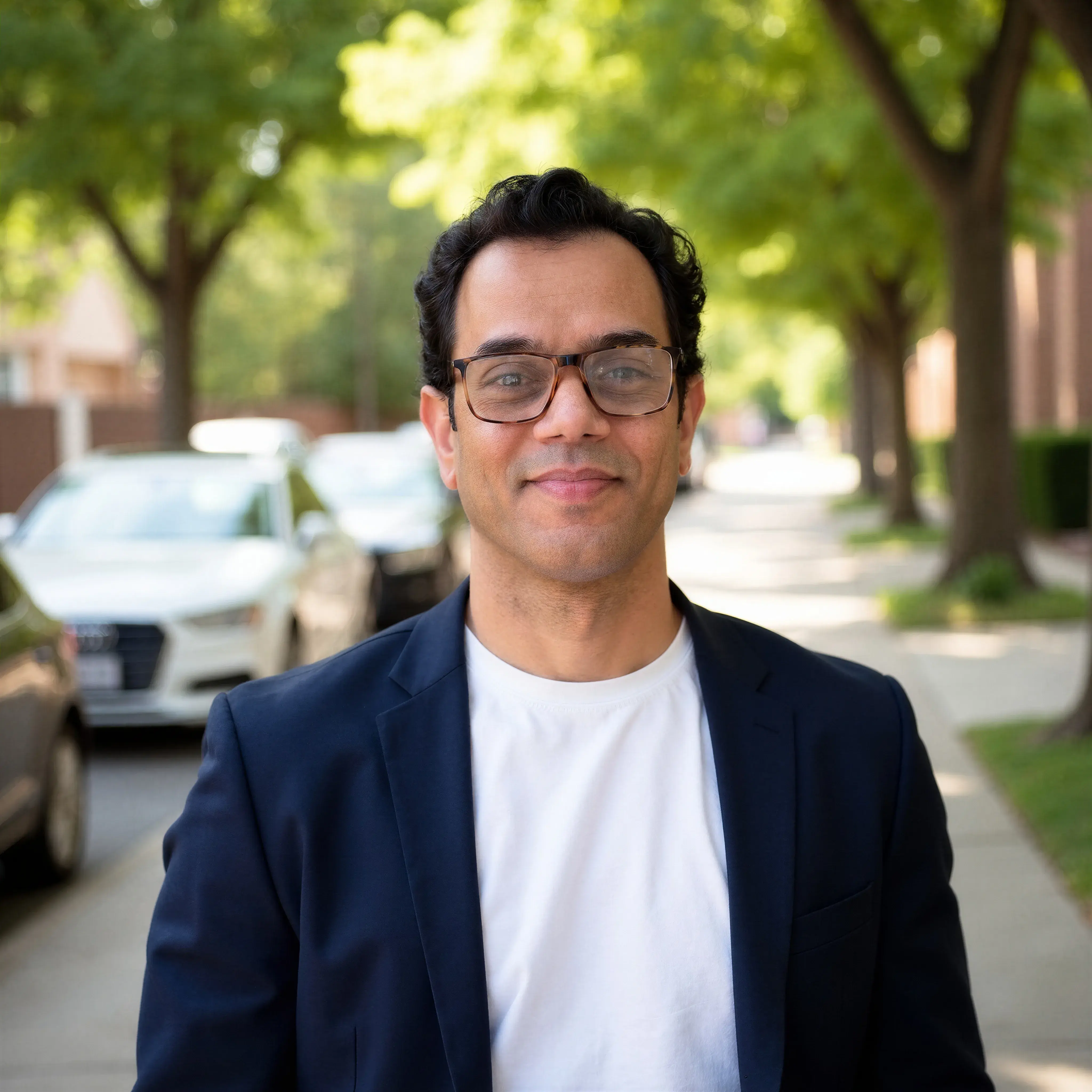 A dark haired man wearing glasses, a white t shirt and a navy blazer smiling pleasantly at the camera