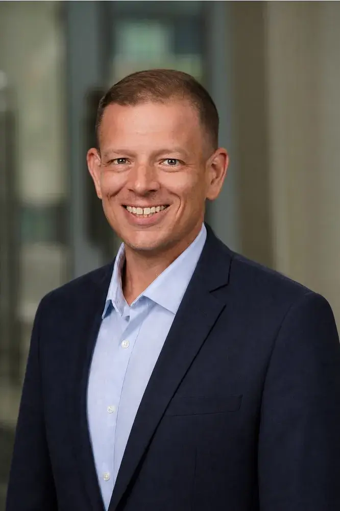 A man in a blue button down and navy jacket smiles at the camera 